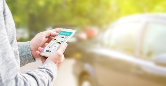 close up of man holding a phone looking at a rideshare application, standing in front of a small car